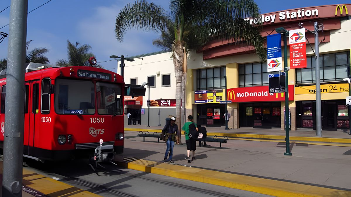 A red MTS trolley parked at the San Ysidro Trolley Station near local commercial buildings and pedestrians.