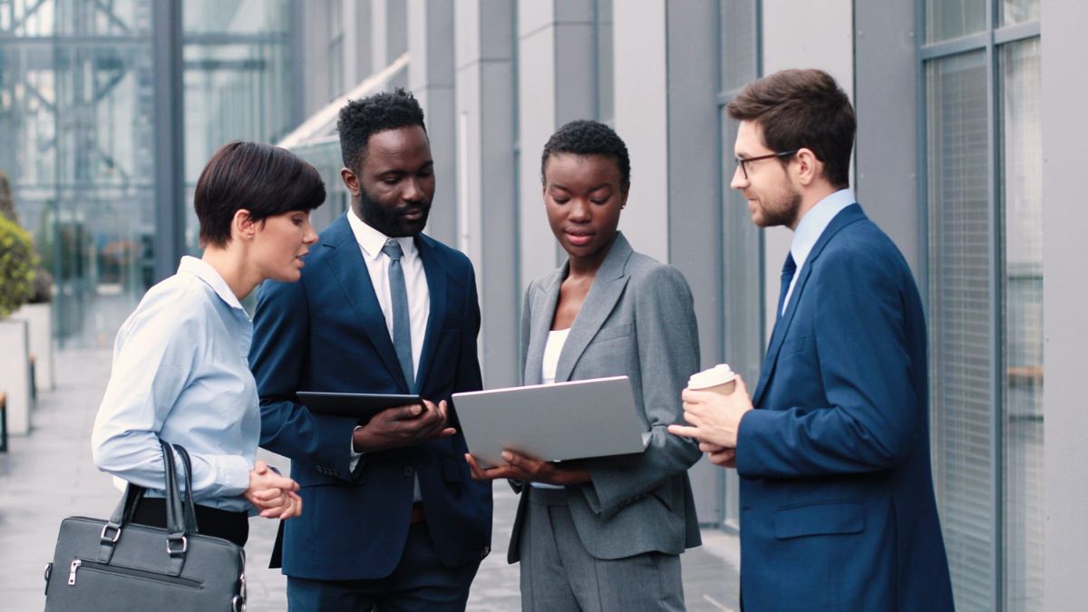 Four diverse business professionals in formal suits reviewing sales data on a laptop and tablet outside a corporate building.