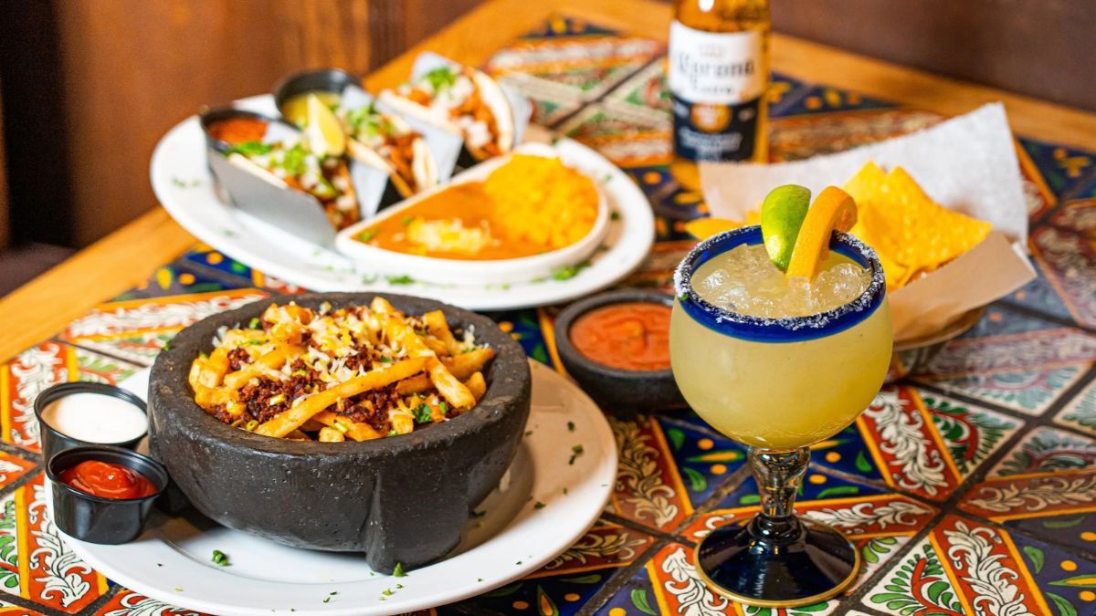 A table spread featuring Mexican dishes including loaded fries in a stone bowl, tacos, chips, salsa, and a margarita