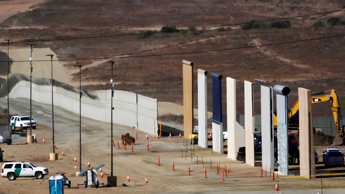 The US-Mexico border wall extending across an arid landscape with security vehicles and safety cones.