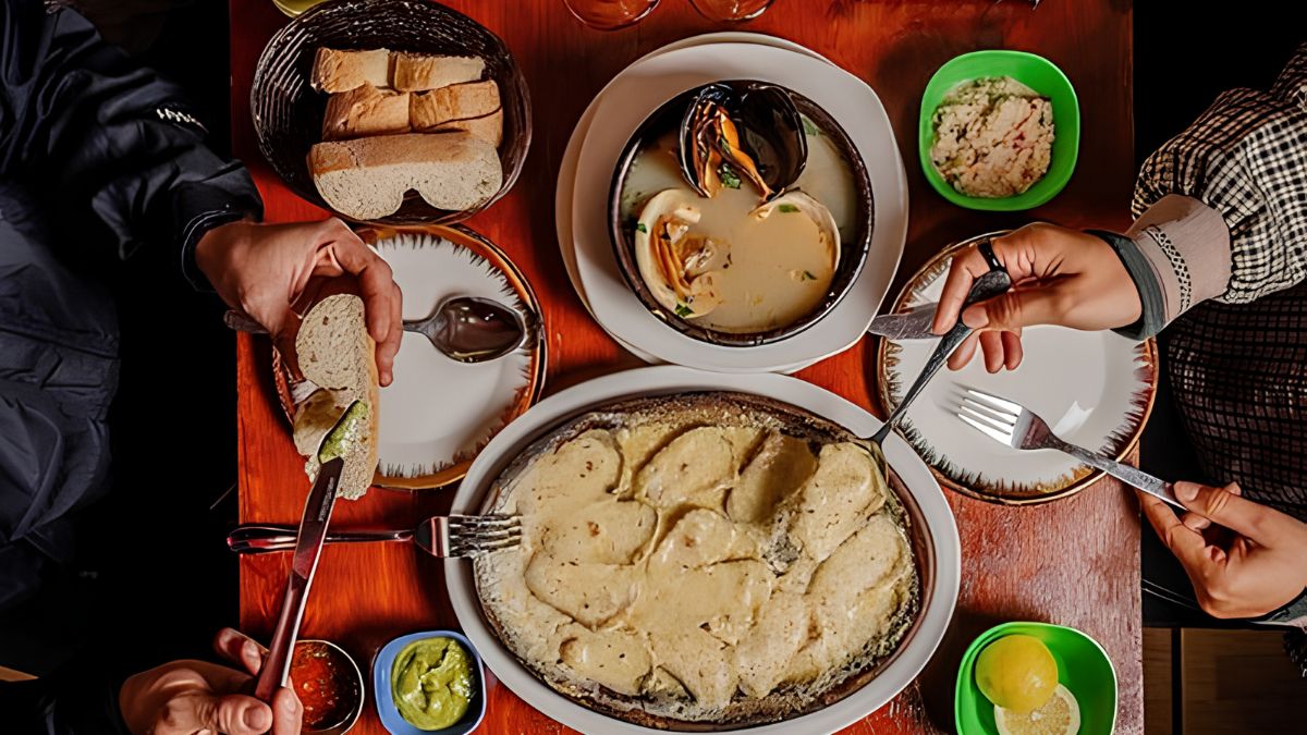 An overhead photograph captures a dining table laden with food and people. In the center, a large oval platter holds creamy, scalloped-looking potatoes. Above it, a dark ceramic bowl is filled with steamed mussels in a broth. To the left is a basket of sliced bread, and small bowls contain various sauces like guacamole. Two people, one in a black jacket and one in a plaid shirt, are seen from the chest up, with their hands using cutlery and bread.