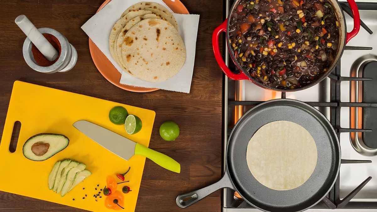 An overhead view of a kitchen counter with a plate of tortillas, a pot of bean stew, sliced avocado on a yellow cutting board, and a tortilla warming in a cast-iron skillet.