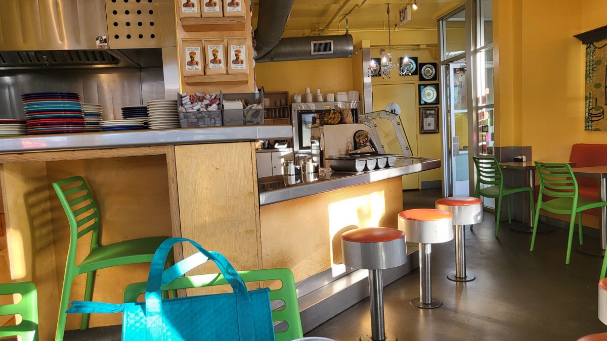 The warm interior of a local café featuring a wooden counter, diner-style counter stools with red seats, green chairs, and shelves stocked with coffee supplies.