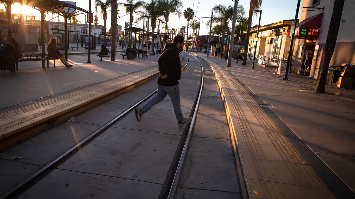 A man in a dark jacket and jeans is captured mid-stride, running across the parallel steel rails of a trolley track system. He moves from left to right across the frame. The scene is set in an urban outdoor transit station or street environment. Palm trees and modern buildings line the background. The scene is strongly illuminated by the warm, low-angle golden light of the setting sun, casting long shadows and creating bright highlights. Other people are visible on the platform in the distance.
