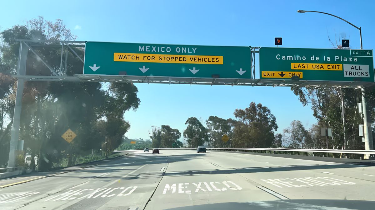 A first-person perspective photograph taken from a moving vehicle on a multi-lane highway, approaching a large green overhead highway sign. The sign on the left lane directs to "MEXICO ONLY" with an additional warning "WATCH FOR STOPPED VEHICLES." The sign on the right lane indicates "EXIT 1A" for "Camino de la Plaza" and designates it as the "LAST USA EXIT" for "ALL TRUCKS." Pavement markings also clearly indicate "MEXICO" in large white letters on the road. The background shows a tree-lined highway leading towards the border under a clear blue sky.
