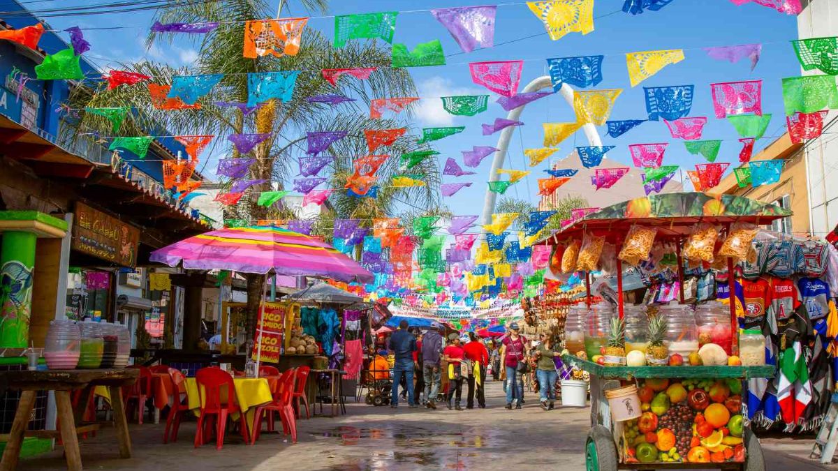 A colorful outdoor street market decorated with traditional papel picado banners, food carts, and shoppers