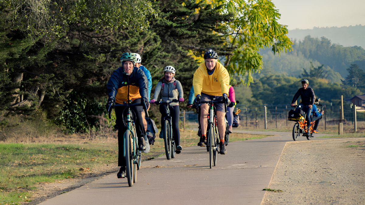 A group of cyclists riding bikes along an outdoor paved path surrounded by trees