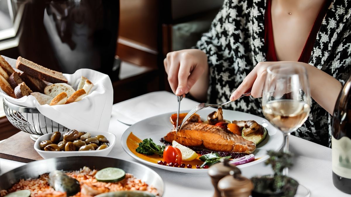 A person cutting a gourmet salmon dish at a restaurant table with bread and wine