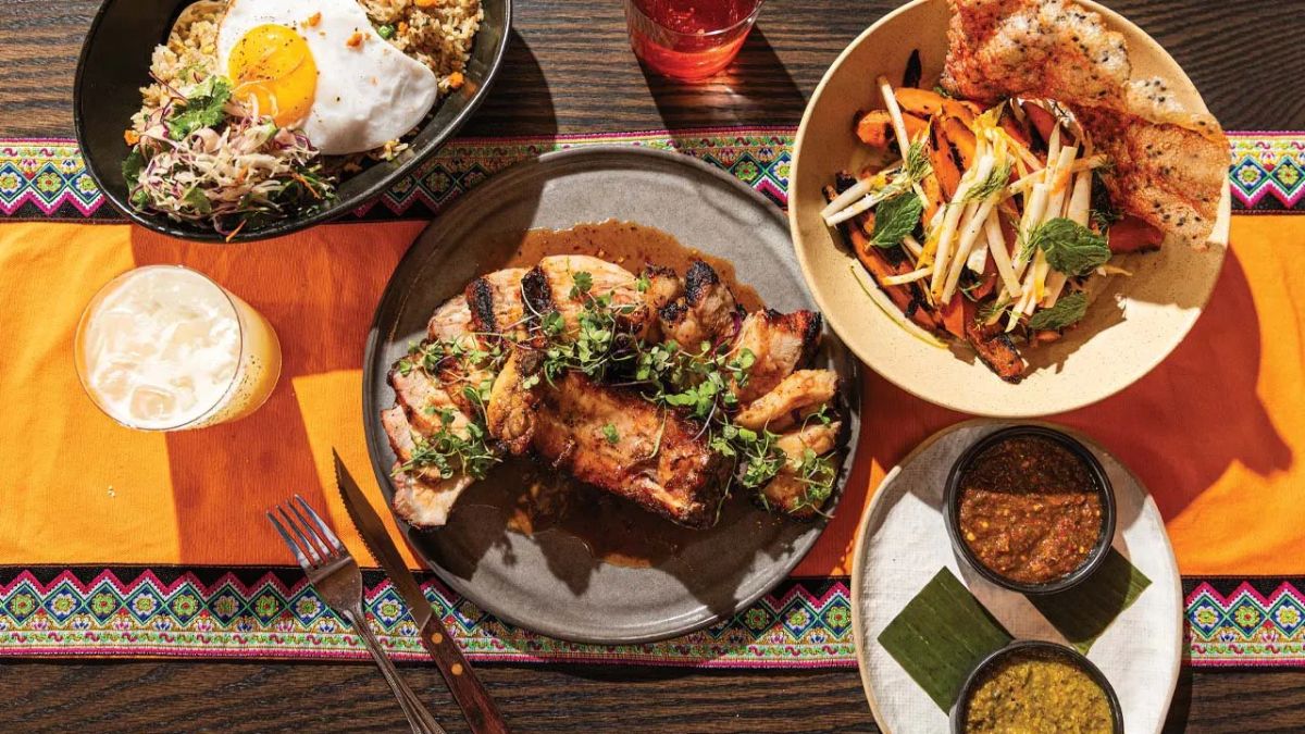 An overhead shot of a table spread with various dishes, including grilled sliced meat garnished with microgreens, a bowl of rice topped with a fried egg, a side of roasted root vegetables, two small bowls of dipping sauces, and glasses of drinks, all set on a bright orange patterned runner.