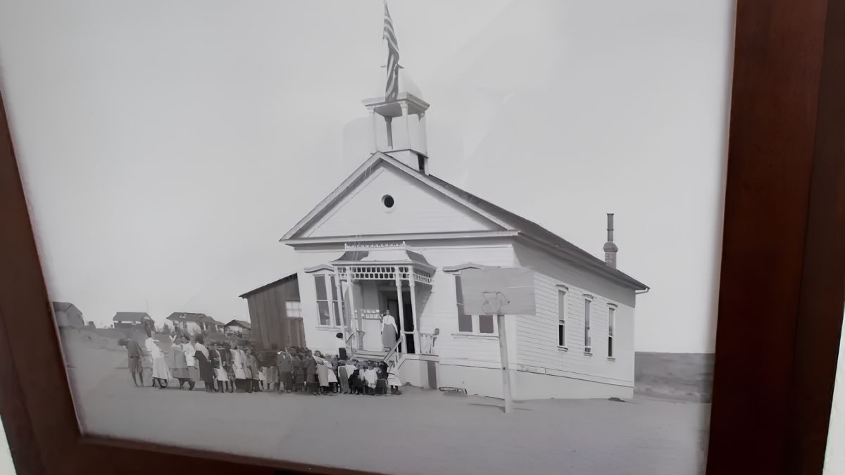 A framed vintage black-and-white photograph showing a historic school building with a bell tower, flag, and a group of children gathered outside.