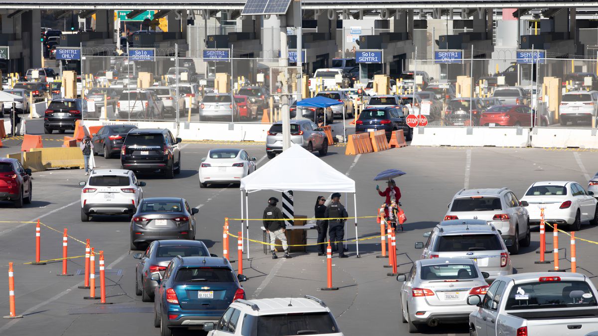 A daytime photograph of the busy vehicle lanes at the San Ysidro Port of Entry, with long queues of cars and multiple inspection booths. Security personnel stand under a white tent.