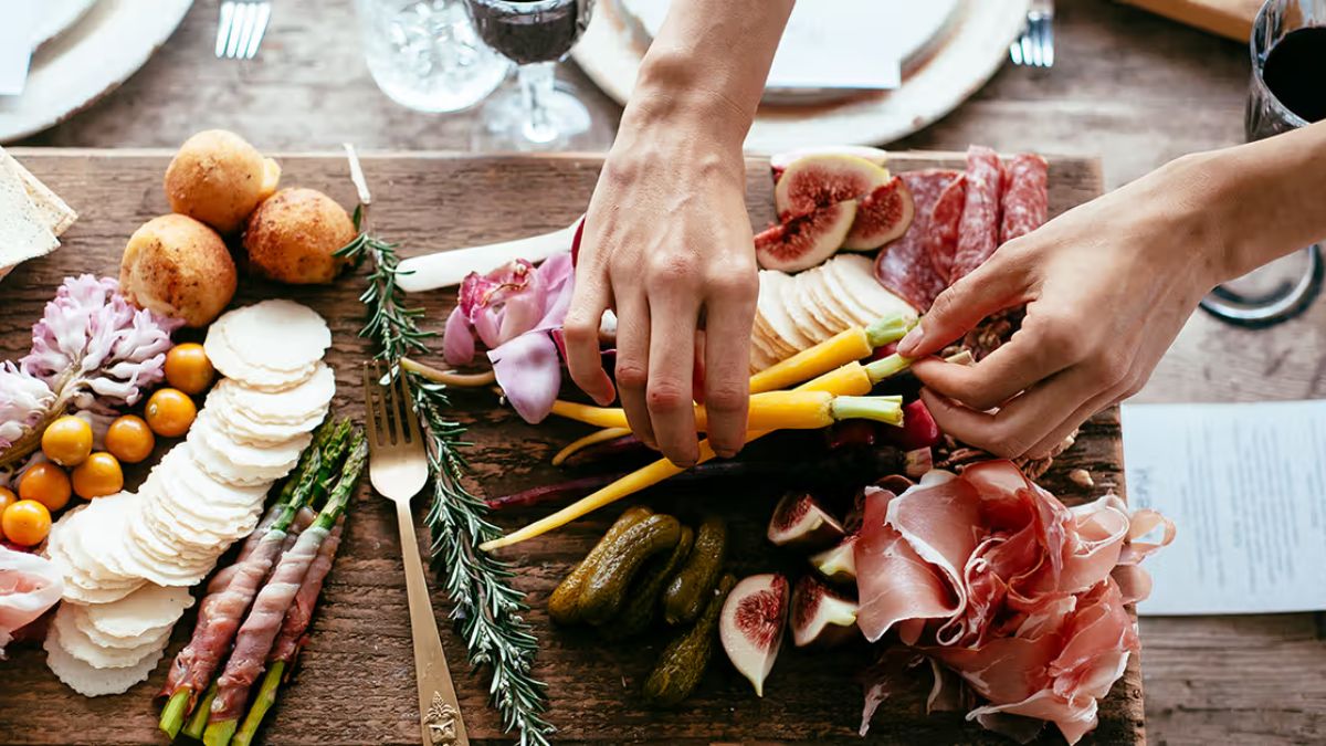 An overhead, close-up photograph of an abundant charcuterie board on a rustic wooden table. Two hands are in the center, actively arranging small yellow carrots and figs. The board is generously filled with sliced meats (prosciutto, salami), fresh purple and yellow cherry tomatoes, asparagus wrapped in meat, gherkin pickles, crackers, cheese, and fresh purple flowers for garnish. A vintage gold fork rests on the left. Blurred wine glasses are in the background.