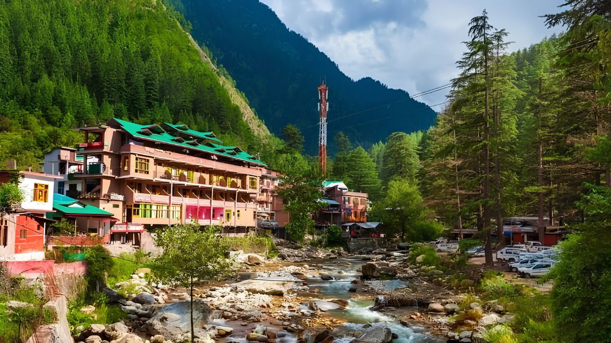 A scenic village nestled alongside a flowing river with lush green mountains and tall trees in the background.