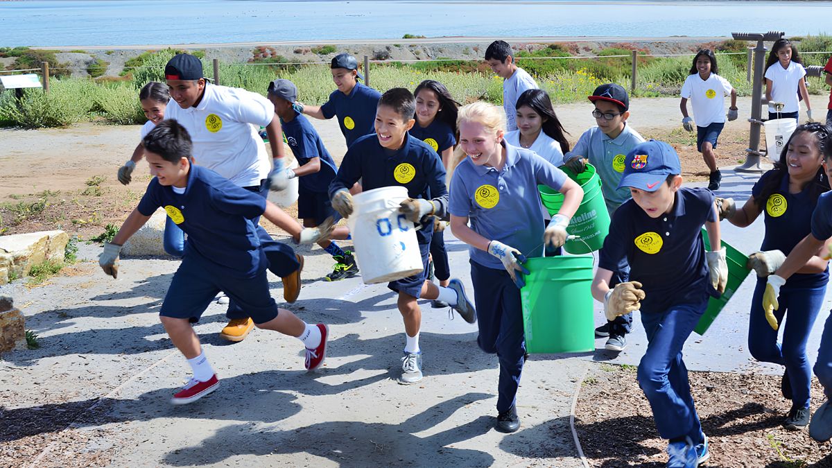 A group of energetic young volunteers and children carrying buckets and wearing work gloves, participating in a neighborhood improvement project in San Ysidro.