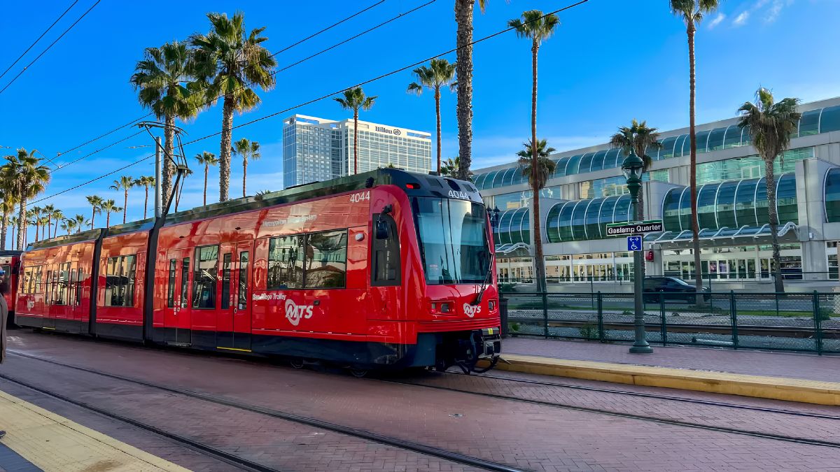 A red San Diego MTS trolley train stopped at a station platform with palm trees and buildings in the background.