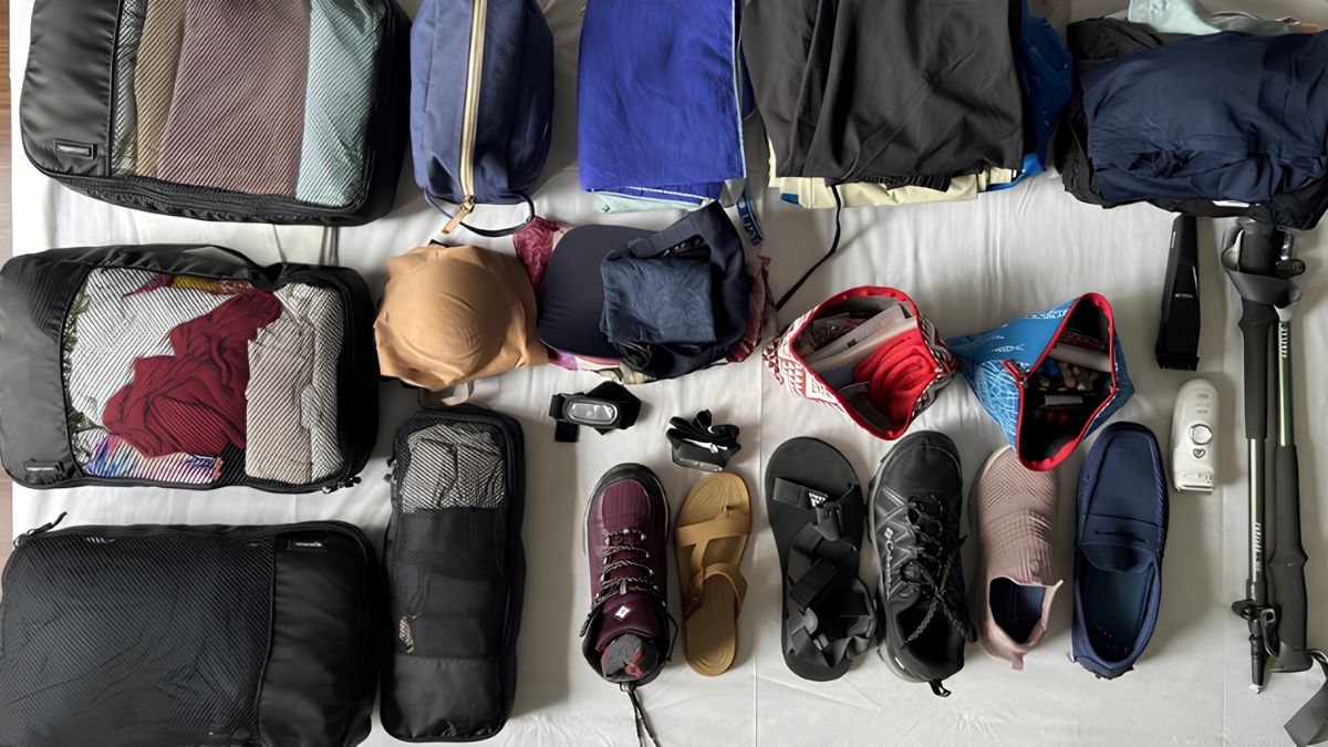 A top-down flat lay view of travel gear laid out on a white surface, including packing cubes filled with clothing, various pairs of shoes and sandals, small accessory pouches, and trekking poles.