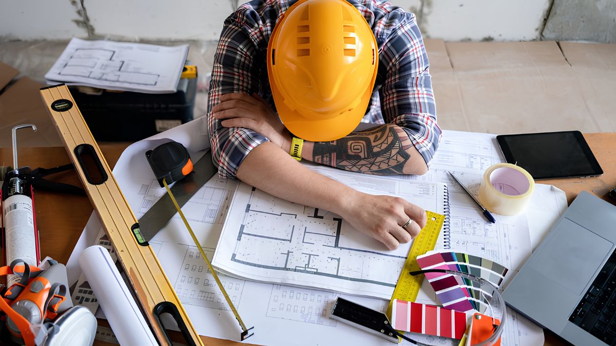 A construction worker wearing a yellow hard hat resting on a desk covered with architectural blueprints, measuring tape, color swatches, and tools.