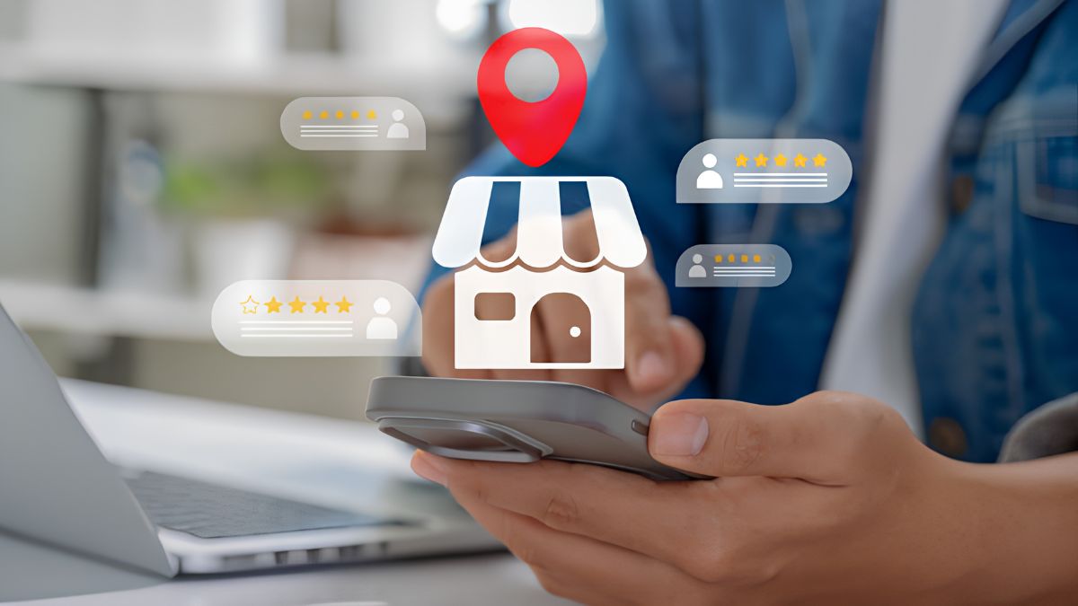A close-up of a person in a denim shirt holding a smartphone. A digital icon of a storefront with a red location pin above it is floating above the phone. Several semi-transparent digital review boxes, showing avatars and yellow star ratings (some four, some five stars), surround the storefront icon. A laptop is visible in the blurred background.