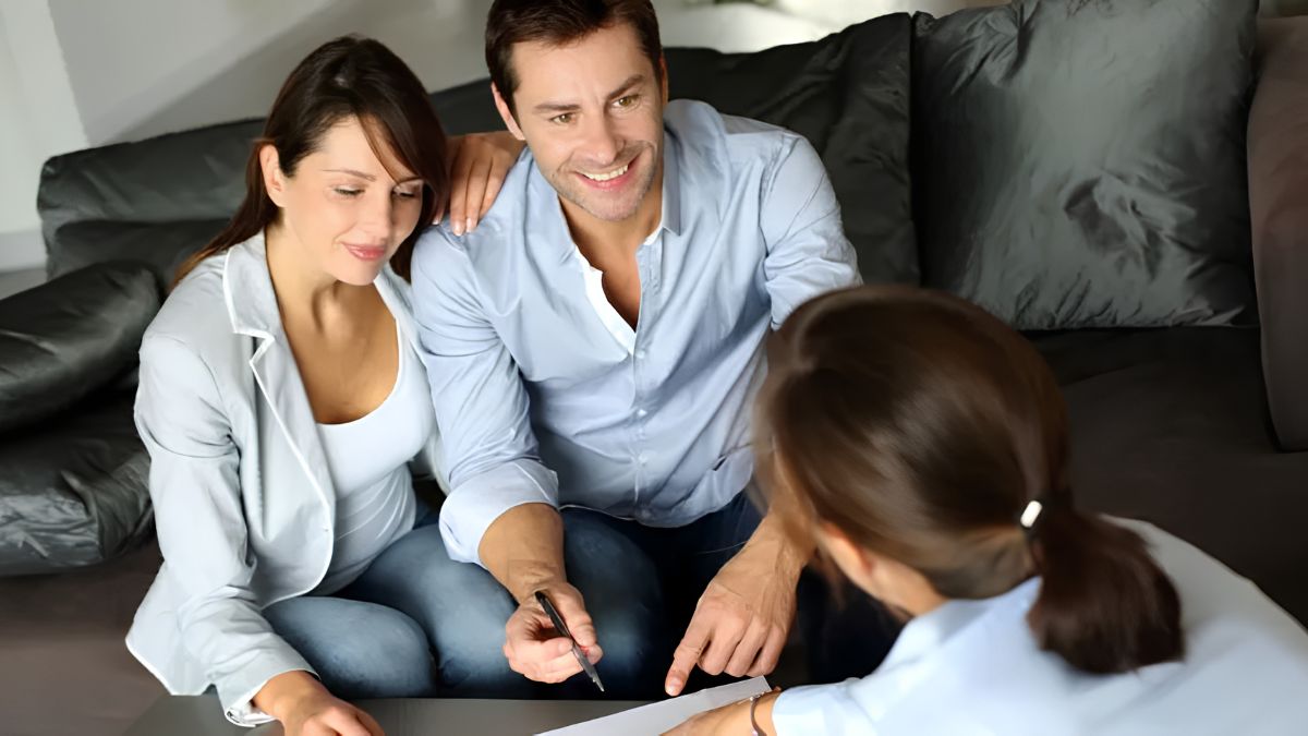 A smiling couple sits together on a dark grey sofa, consulting with a female financial advisor whose back is to the camera. The man holds a pen and points at paperwork on a coffee table, while the woman looks on with a pleasant expression.