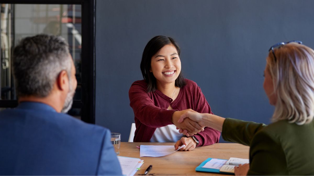 A smiling new employee shaking hands with a manager across a desk during an onboarding meeting in a small business office.
