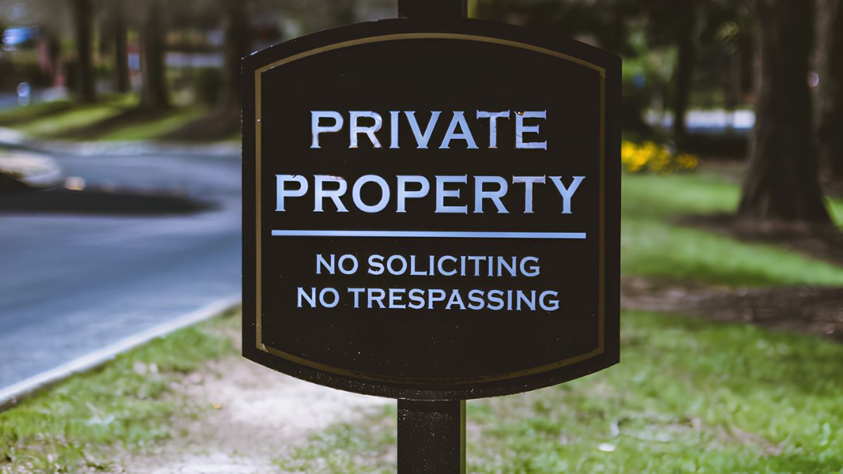 A dark sign reading "Private Property No Soliciting No Trespassing" posted outdoors near a road