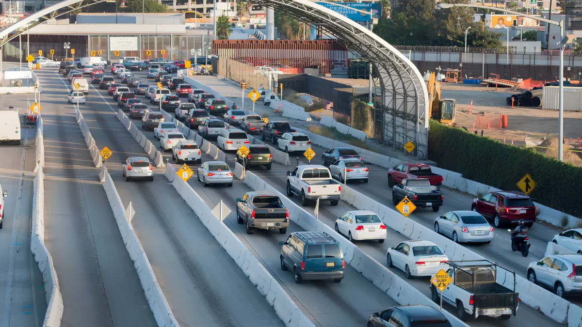 An elevated, high-angle view of a multi-lane roadway packed with cars, trucks, and vans moving in heavy traffic toward a security checkpoint or border crossing marked by concrete barriers and overhead structures.