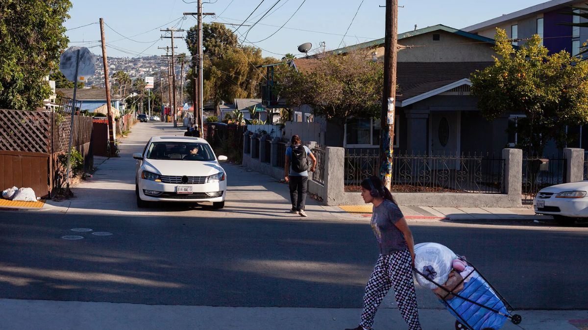 A street view in a residential neighborhood in San Ysidro, showing a person walking with a cart, a car driving down the road, and homes along the sidewalk.