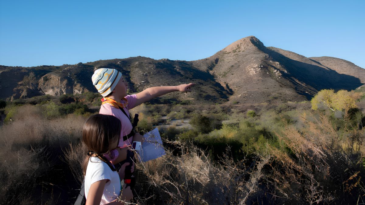 Two people standing outdoors in a natural landscape, with one person pointing toward rolling hills and a mountain peak.