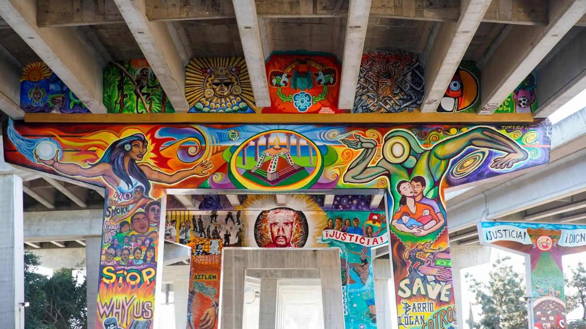 A low-angle photograph shows a large, colorful mural covering the concrete pillars and beams supporting a highway overpass. The mural depicts a complex array of figures, including a central sun face, a woman holding a glowing orb, a couple, and abstract patterns in bright, bold colors like yellows, reds, and blues. Text is integrated into the art, reading "STOP WAR" on one pillar and "SAVE BARRIO LOGAN" with "JUSTICIA!" and "¡DIGNIDAD!" on others. The concrete beams above also feature smaller, distinct painted panels.