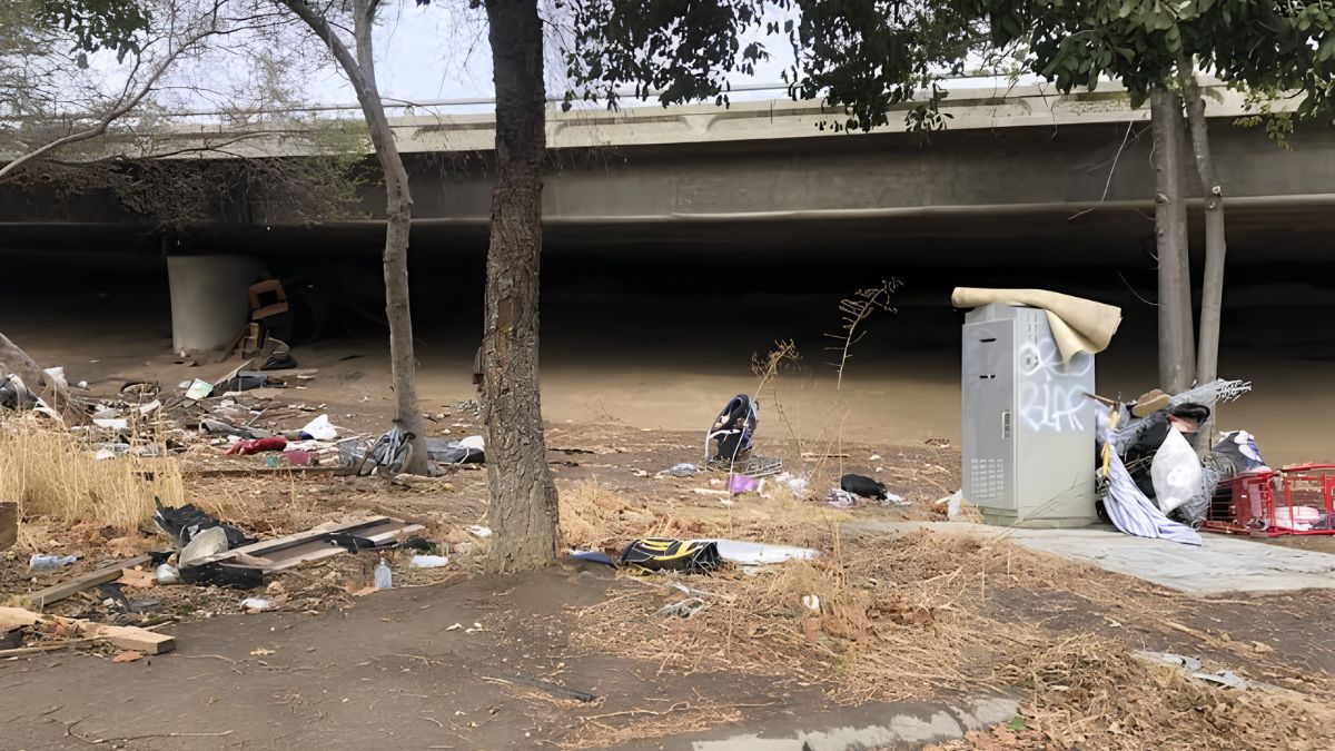 An area under an overpass littered with debris, trash, and graffiti on a utility box