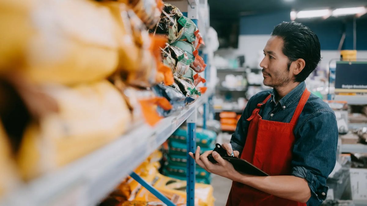 Retail employee in a red apron reviewing stock on store shelves using a clipboard