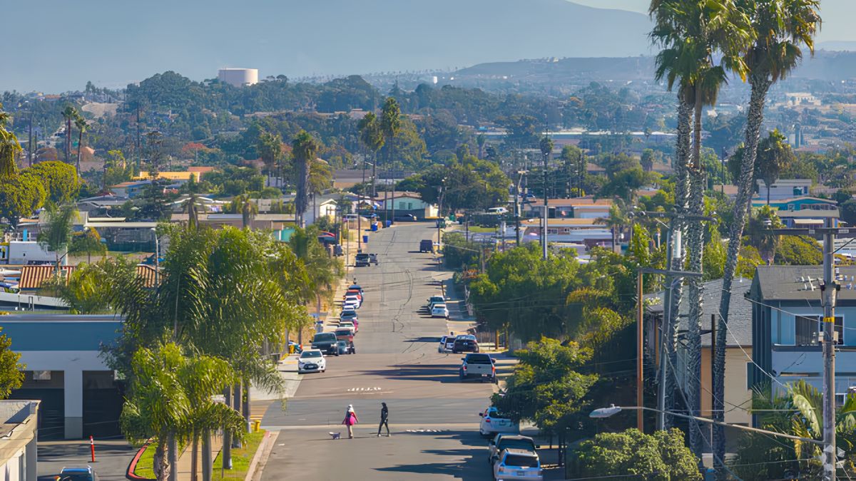 A photograph taken from an elevated position looks down a long, straight residential street that descends a steep hill in San Diego, California. Palm trees and utility poles line both sides of the street, which is flanked by houses and apartment buildings. Cars are parked along the curbs, and a few are driving in both directions. In the distance, past the end of the street, a valley is visible with more urban development, leading up to green, tree-covered hills under a clear blue sky. Two people are walking on the sidewalk in the foreground.