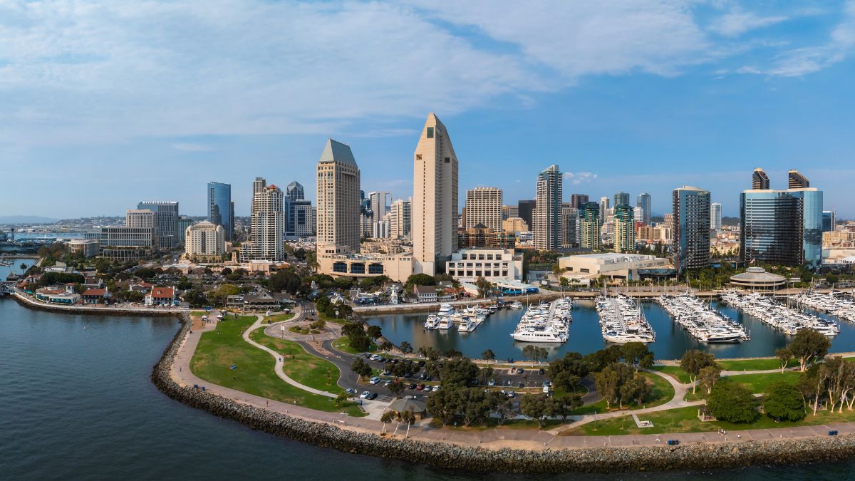 Panoramic aerial view of the San Diego city skyline, modern skyscrapers, and a docked marina with boats along the waterfront.