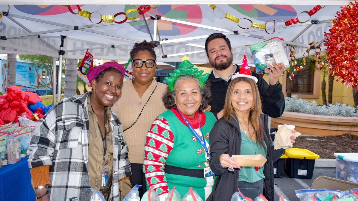 A group of diverse volunteers and community workers smiling together under a decorated charity tent with donation packages.