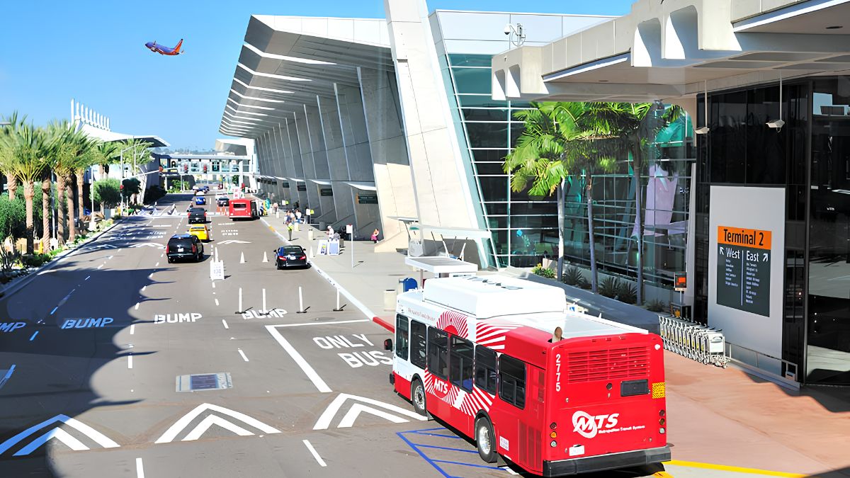 An elevated daytime photograph capturing the busy curbside of San Diego Airport's Terminal 2. In the foreground, a red and white Metropolitan Transit System (MTS) airport shuttle bus is pulling away from the curb, marked with "ONLY BUS" on the asphalt. Palm trees line the left side of the multi-lane road. The modern glass and concrete terminal building is on the right, with clear signage for "Terminal 2 West | East." In the upper left sky, a Southwest Airlines jet is in flight. Cars are visible in the traffic lanes, and pedestrians are waiting at the curb.