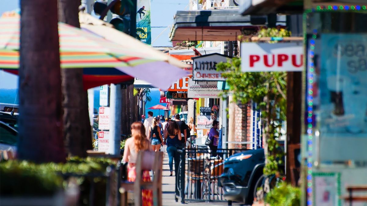 A sunlit, daytime photograph looking down a bustling sidewalk in La Jolla, California. In the foreground, a woman in a white top and patterned skirt with long reddish hair walks away from the camera. The sidewalk is crowded with pedestrians walking towards the distant blue ocean. Numerous colorful storefronts and restaurants line the path, featuring various signs. Prominent signs include a red and white sign reading "PUPU" on the right, and partially obscured signs for "LITTLE CHINESE CAFE" and "BEACH" further down. A black car is parked on the right. The atmosphere is lively and warm.
