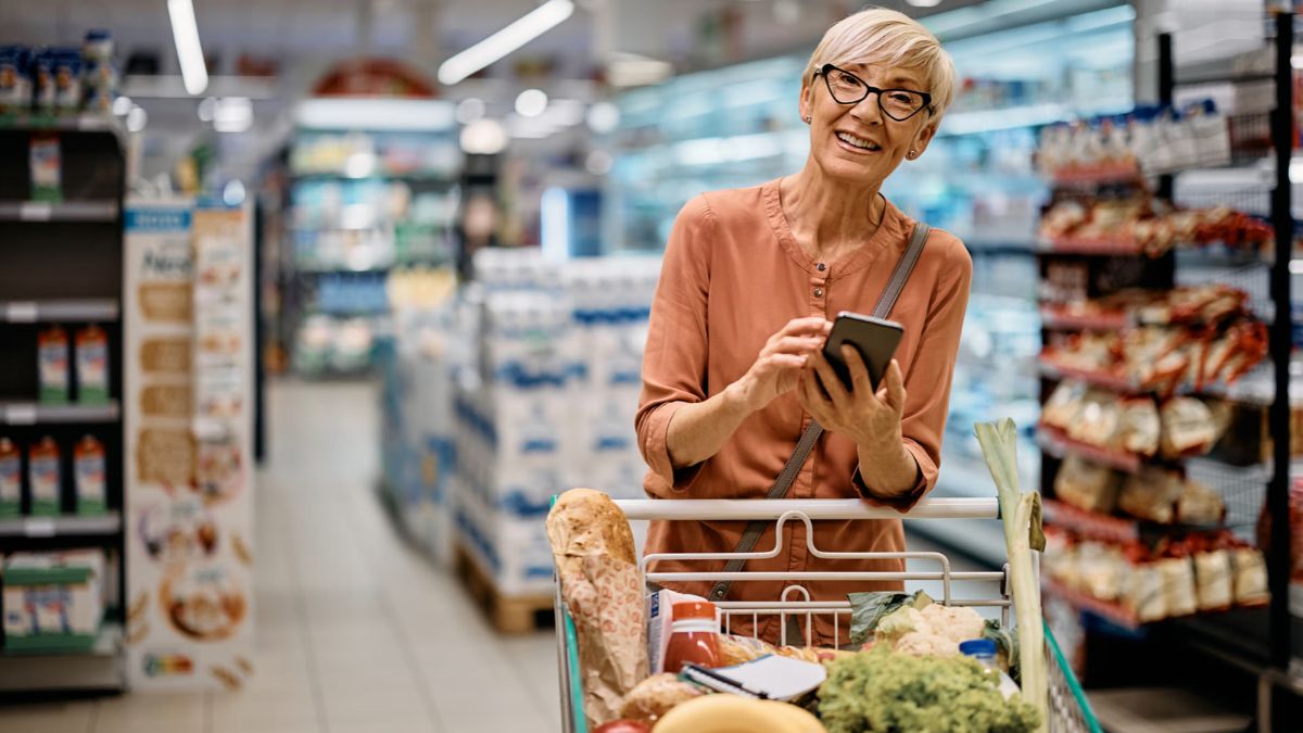Smiling senior woman holding a smartphone while shopping with a grocery cart in a supermarket aisle
