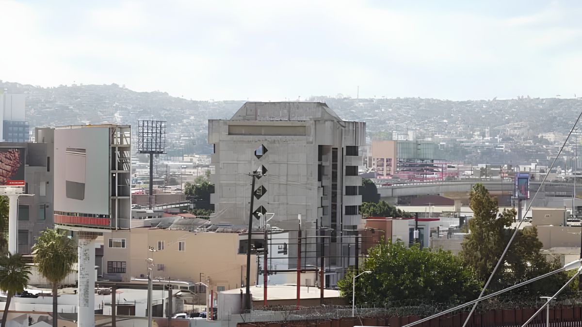A daytime, medium-angle photograph captures a view of a city in a valley, likely Tijuana, Mexico, taken from the US side of the border. In the foreground, parts of the metal border fence with razor wire are visible. The city features a mix of low-rise and mid-rise concrete and stucco buildings, with a prominent, aged brutalist-style office building in the center. Large, blank billboards are on the left, and a communications tower stands behind them. Rolling, hazy hills with more development rise in the background under a pale, overcast sky.