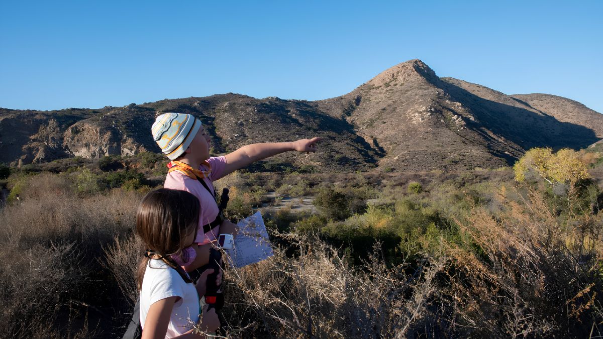 Two children hiking outdoors on a sunny day, with one child pointing towards a large mountain peak in the background.