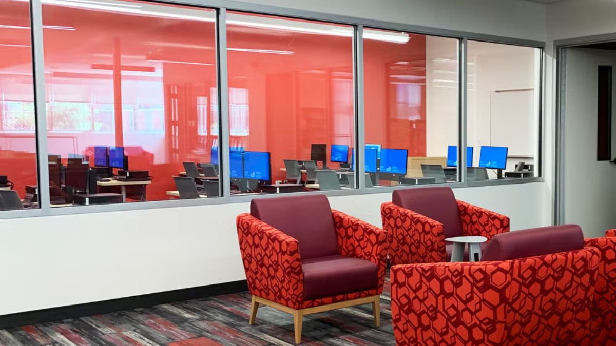 Modern public library interior featuring comfortable red patterned armchairs and a computer lab visible through large glass windows