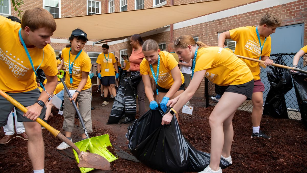 A sunlit outdoor photograph captures a team of six young adults, all wearing matching yellow "LEADERSHIP IN ACTION" t-shirts and blue lanyards, actively participating in a mulching project. In the foreground left, a man holds a yellow-handled shovel. Next to him, a woman in a visor holds a light green shovel. In the center, two women in gloves carefully hold open a large black trash bag as another volunteer shovels mulch into it. On the far right, a man in a yellow shirt is also shoveling mulch. They are standing on a bed of dark brown wood chip mulch. The background features a brick building, a black chain-link fence, and other blurred volunteers under a large beige shade sail. The atmosphere is collaborative and focused.