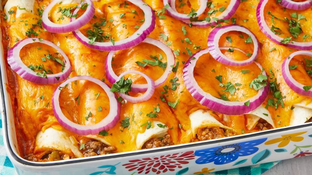 A close-up view of a baked casserole dish filled with savory enchiladas covered in melted cheese, red onion rings, and fresh cilantro