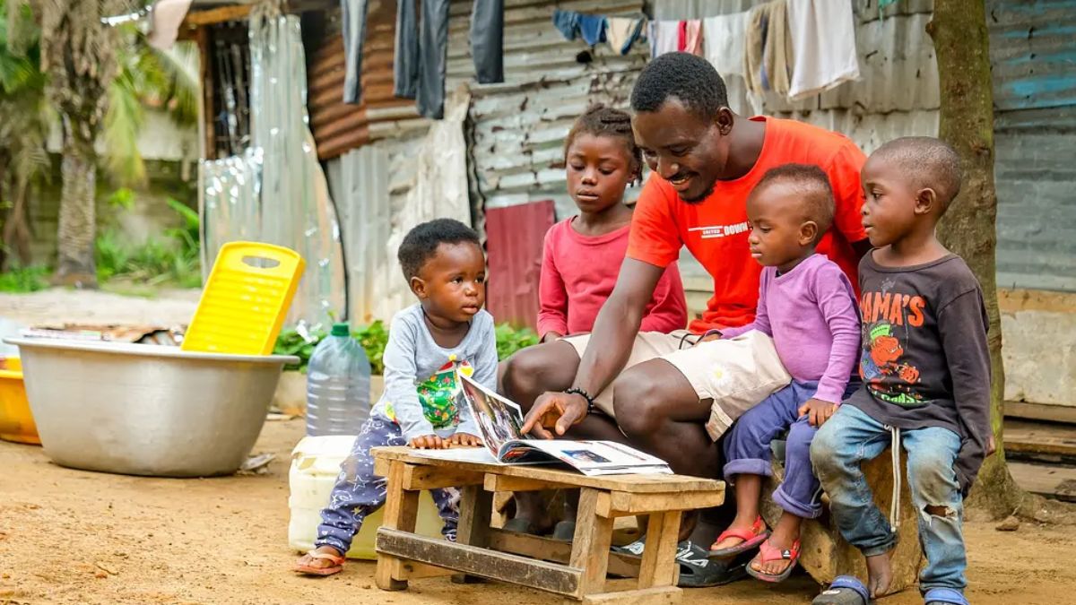 An adult man in a red t-shirt sits outdoors with four young children, pointing to an open photo album or storybook resting on a small wooden stool.