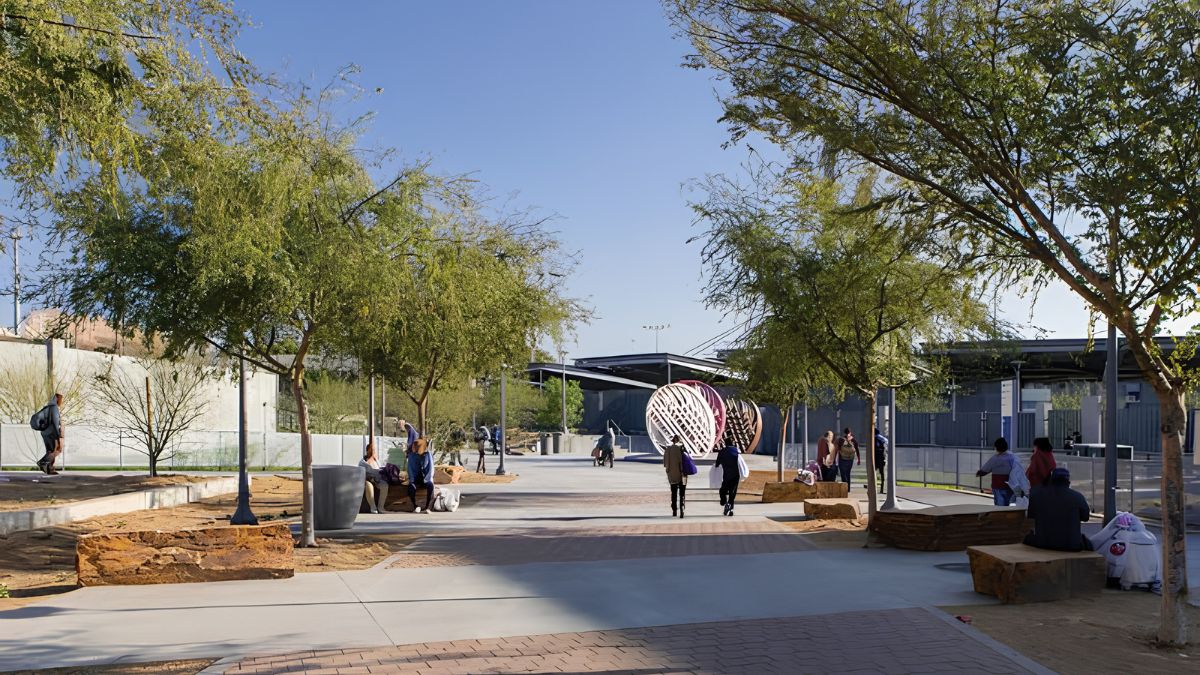 A photograph of a modern outdoor public plaza on a sunny day. A paved walkway leads the eye towards the background, flanked by desert landscaping with acacia trees and large rock benches. Several people are scattered throughout the space, some sitting on the benches and others walking. In the mid-ground, a large, spherical, intertwined metal sculpture serves as a focal point. Beyond the plaza, contemporary buildings with shade structures are visible. The sky is a clear blue.