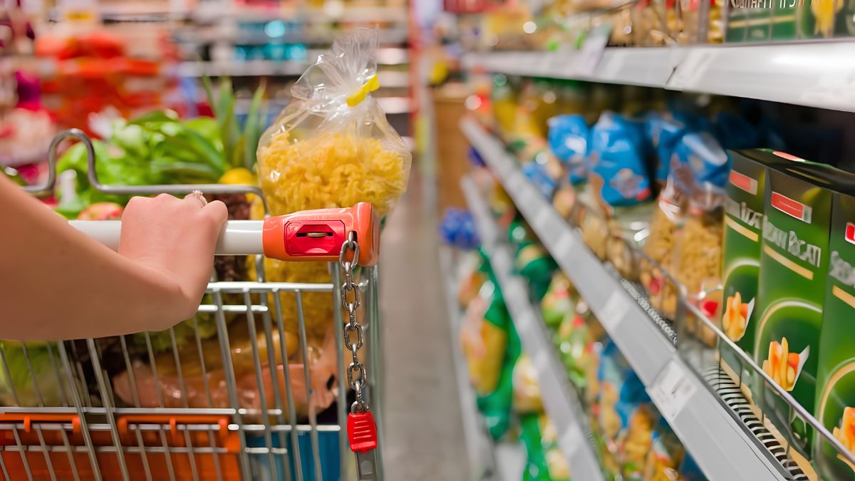A shopper pushing a grocery cart filled with food and pasta down a supermarket aisle lined with stocked shelves.