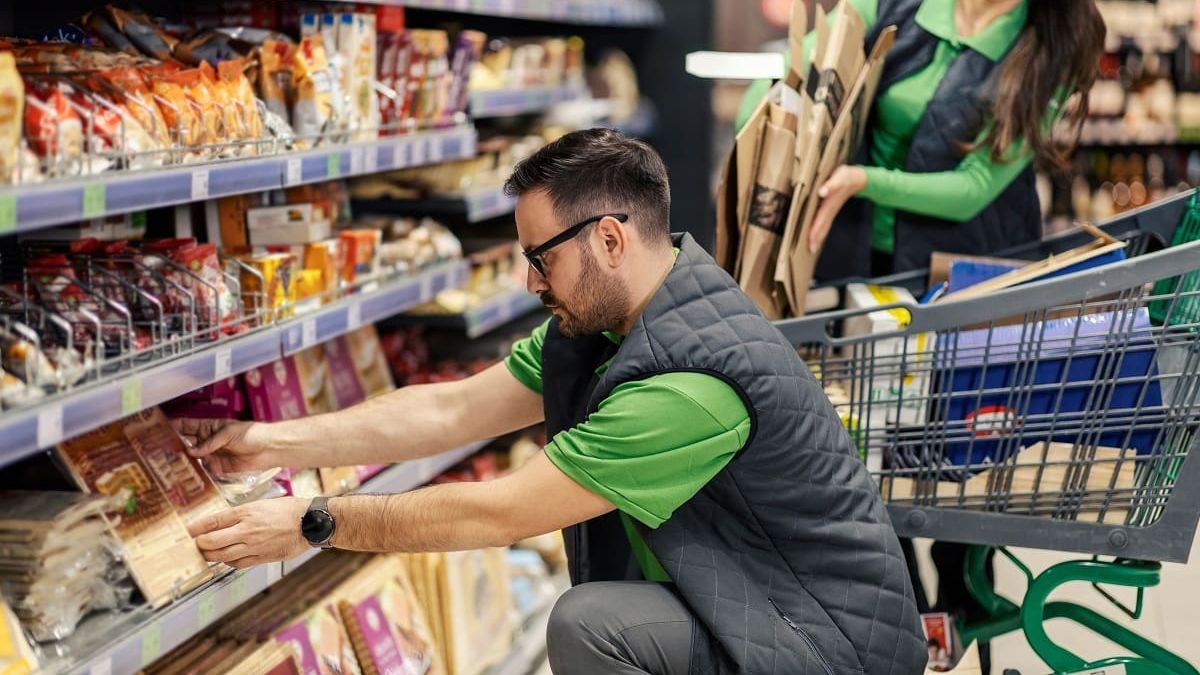 A shopper inspecting packaged food items on a grocery store shelf next to a shopping cart.