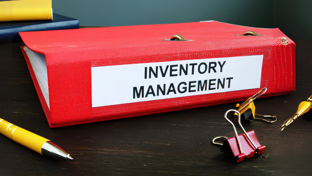 A close-up photograph of a large red office binder lying horizontally on a dark wood surface. A white label on the spine clearly reads "INVENTORY MANAGEMENT" in black, bold, sans-serif font. To the left is a yellow pen. To the right are various metal binder clips (one red and silver, one yellow and silver). A blue and yellow notebook is partially visible in the blurred background.