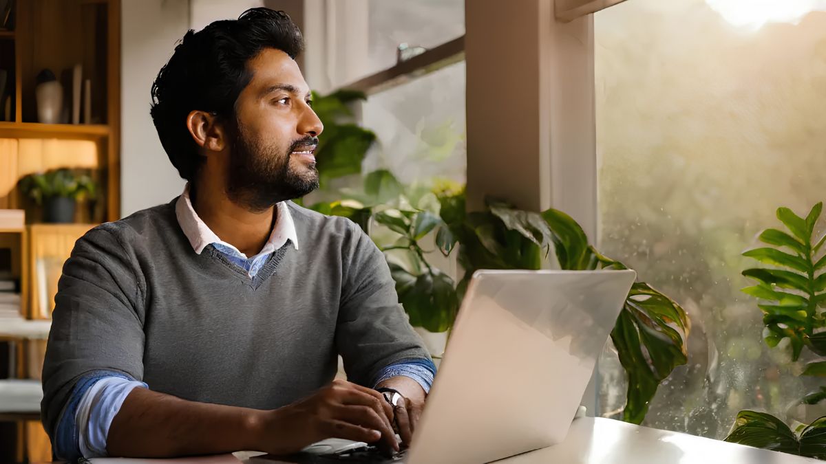A smiling South Asian man in a grey sweater sits at a desk with a laptop, looking out a bright, sunlit window with green houseplants.