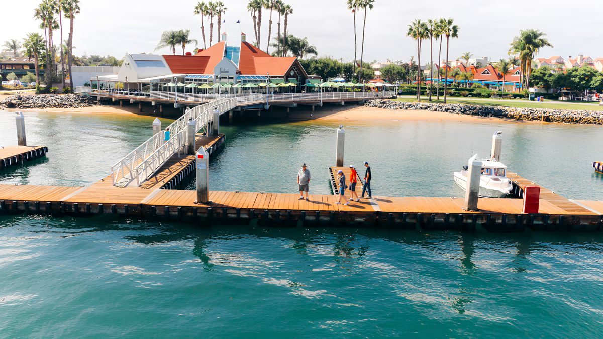 An elevated view of a wooden marina dock on turquoise water. A group of four people walks along the main dock. A metal gangway connects the dock to a waterfront building with a red roof and green umbrellas, surrounded by palm trees. A small white boat is moored nearby.