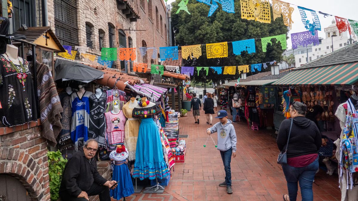 People walking along a historic brick walkway lined with shops selling traditional clothing and crafts, decorated with colorful papel picado banners overhead.